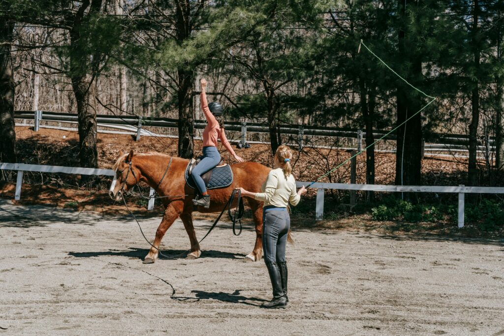 Two women practicing horseback riding in a sunny outdoor arena.